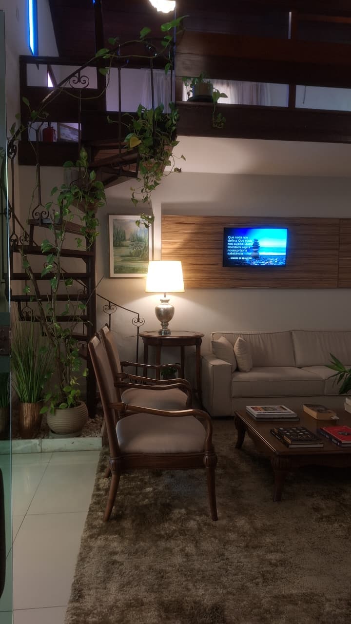 Recepção com sofá claro, poltronas de madeira, plantas e escada em caracol