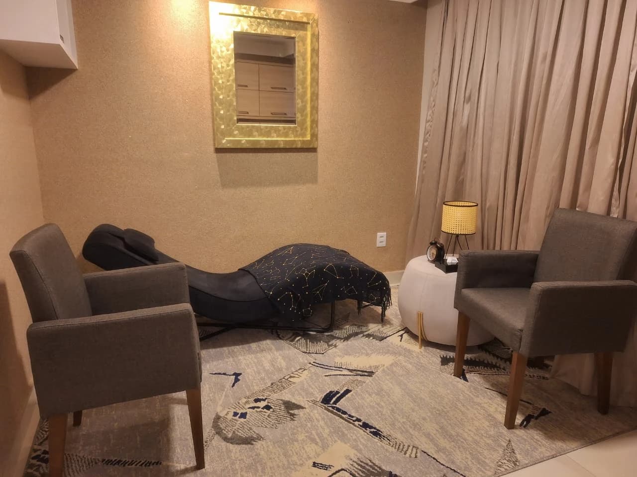 Sala de atendimento com divã, duas poltronas, luminária acesa e espelho dourado